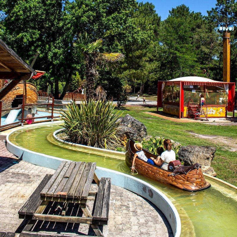 Deux personnes embarquent dans un canoë sur un canal du parc d'attractions Bordeaux Kid Parc, entouré de végétation, de rochers et d’attractions colorées.