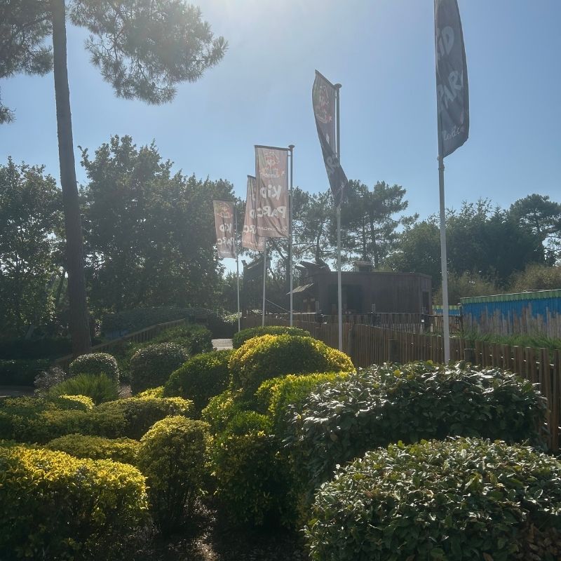 Drapeaux du Kid Parc flottant au vent à l’entrée du parc, entourés de haies et d’arbustes décoratifs sous un ciel ensoleillé.